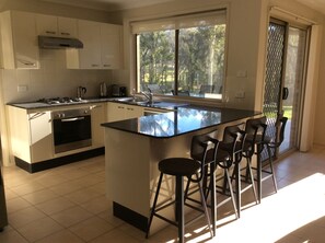 A modern kitchen with a large island, bar stools, and a view of the outdoors through a sliding door. - Idyllic Coastal Farm Stay: Perfect Holiday Home for a Family! (Narooma)