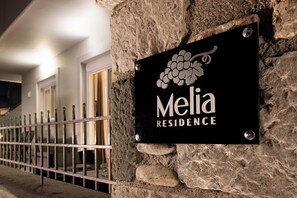 A sign for 'Melia Residence' with a grapevine design, mounted on a stone wall with a metal fence in the background.