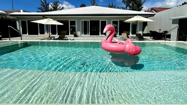Een zwembad met een roze flamingo drijver, ligstoelen, parasols en een gebouw met grote ramen op de achtergrond.
