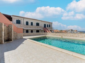 A poolside area with a stone pool, a building with multiple windows, and a clear sky. - Pool, air Conditioning, Parking (Valledoria)