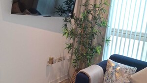 A living room with a dark blue sofa, a large mirror, a potted plant, and a window with blinds. - Central Belfast Apartment/Modern/Ideal for Work & Leisure (Belfast)