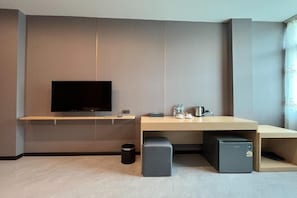 Quarto de hotel moderno com televisão de tela plana, escrivaninha de madeira, pequeno frigorífico e lixeira.