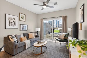 Ein modernes Wohnzimmer mit einem grauen Sofa, einem Couchtisch und einem Fernseher. Ein Balkon bietet Blick auf umliegende Gebäude.