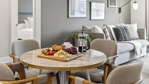 Et rundt spisebord med kaffekande, et fad med frugt og en kop kaffe. Der er to stole omkring bordet. Værelset har en grå sofa, en hvid lænestol og en væg med billeder.