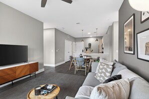 Ein modernes Wohnzimmer mit Flachbildfernseher, einer Holzunterhaltungsecke, einem grauen Sofa, einem Couchtisch und einem Essbereich mit Tisch und Stühlen.