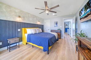 Un dormitorio con cama, mesita de noche, ventilador de techo y vista a la playa a través de una ventana.
