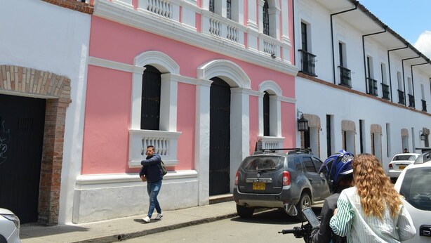 Un edificio rosa y blanco con ventanas arqueadas y balcones, una calle con coches y una persona caminando.