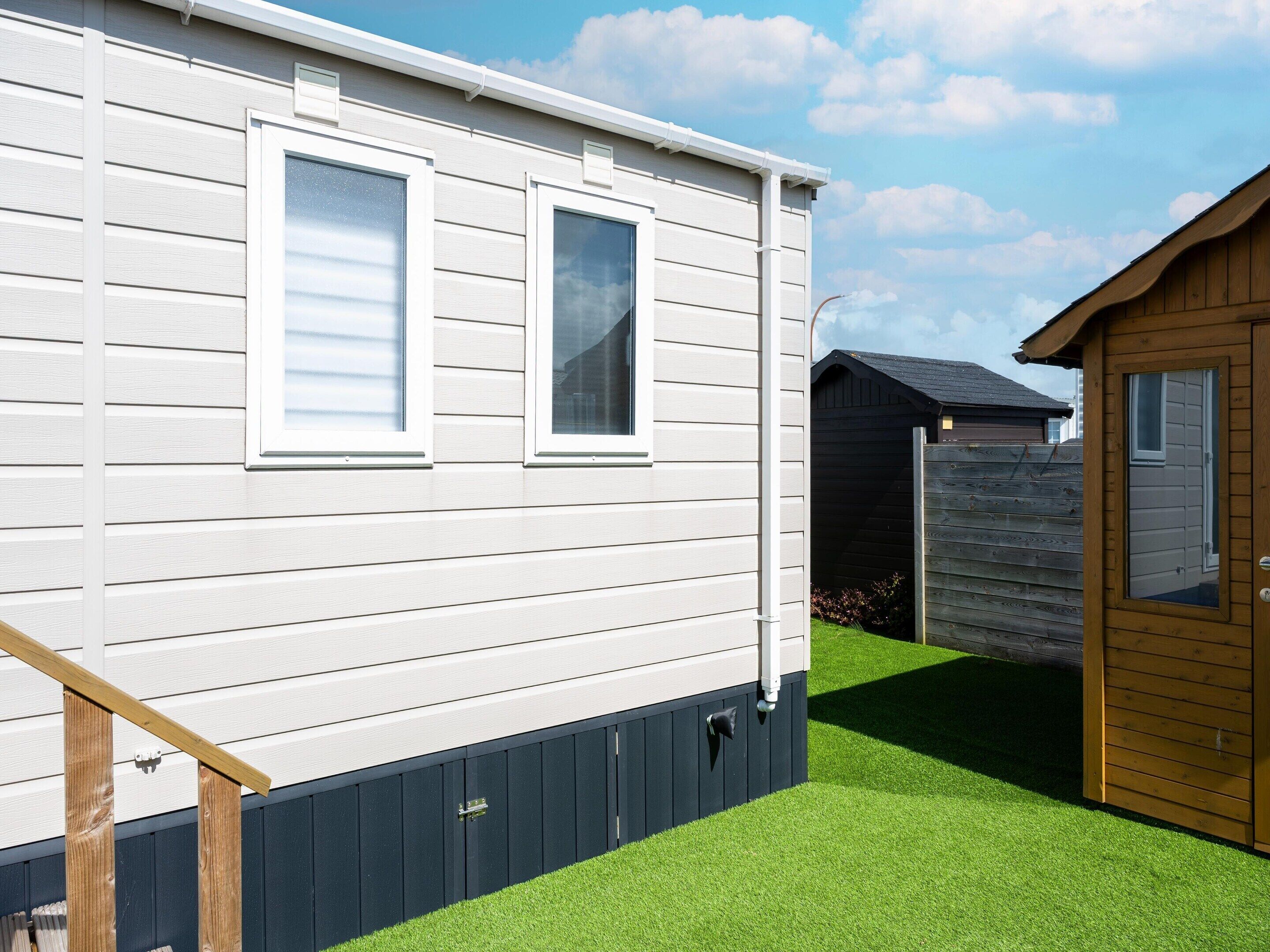 A white mobile home with two windows, a wooden deck, and a small fenced garden area.