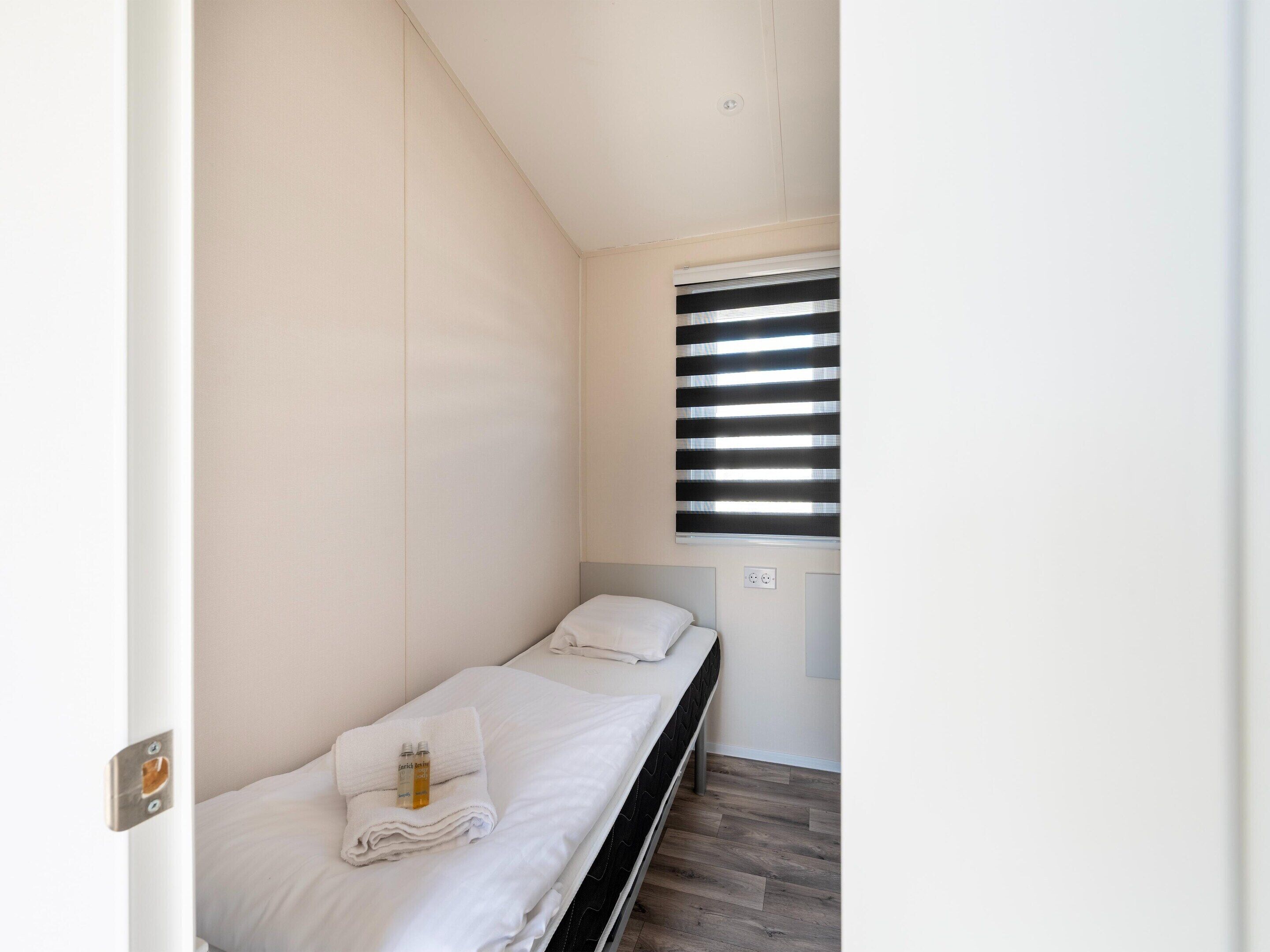 A small, single-bed room with a window, a bedside table, and a wall socket.