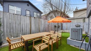 A backyard with a wooden table and chairs, an orange umbrella, a lawn, and a house in the background. - Nebula Ranch: 3BR Space Cowboy Retreat, Fire Pit + Mural (Houston)
