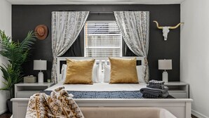 A bedroom with a bed, two bedside tables, a nightstand, a lamp, a large potted plant, a decorative skull, and a window with curtains. - Nebula Ranch: 3BR Space Cowboy Retreat, Fire Pit + Mural (Houston)