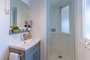 A bathroom with a glass-enclosed shower, a white sink with a wooden cabinet below, a mirror above the sink, and a shelf with a plant and a small vase. - Vigna Sul Mar Family Collection (Comacchio)