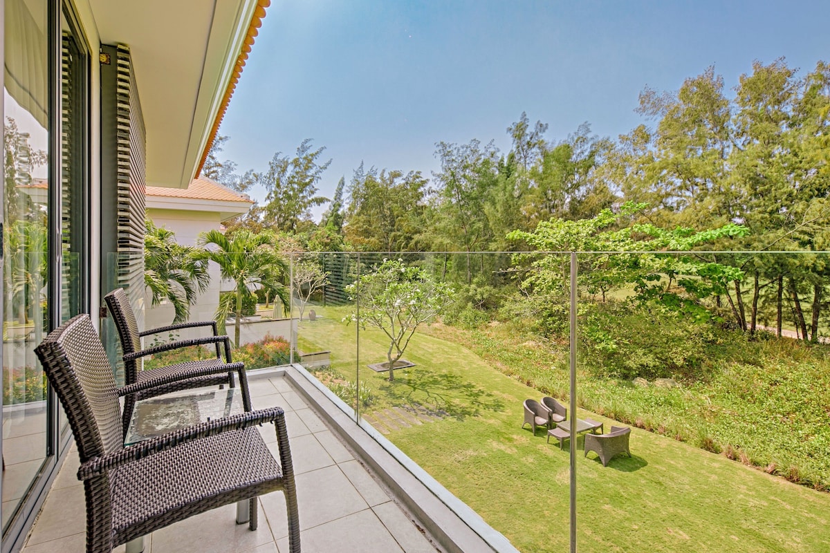 Ein Balkon mit Korbstühlen, einem Glastisch und Blick auf einen gepflegten Garten mit Bäumen und einer Hecke.