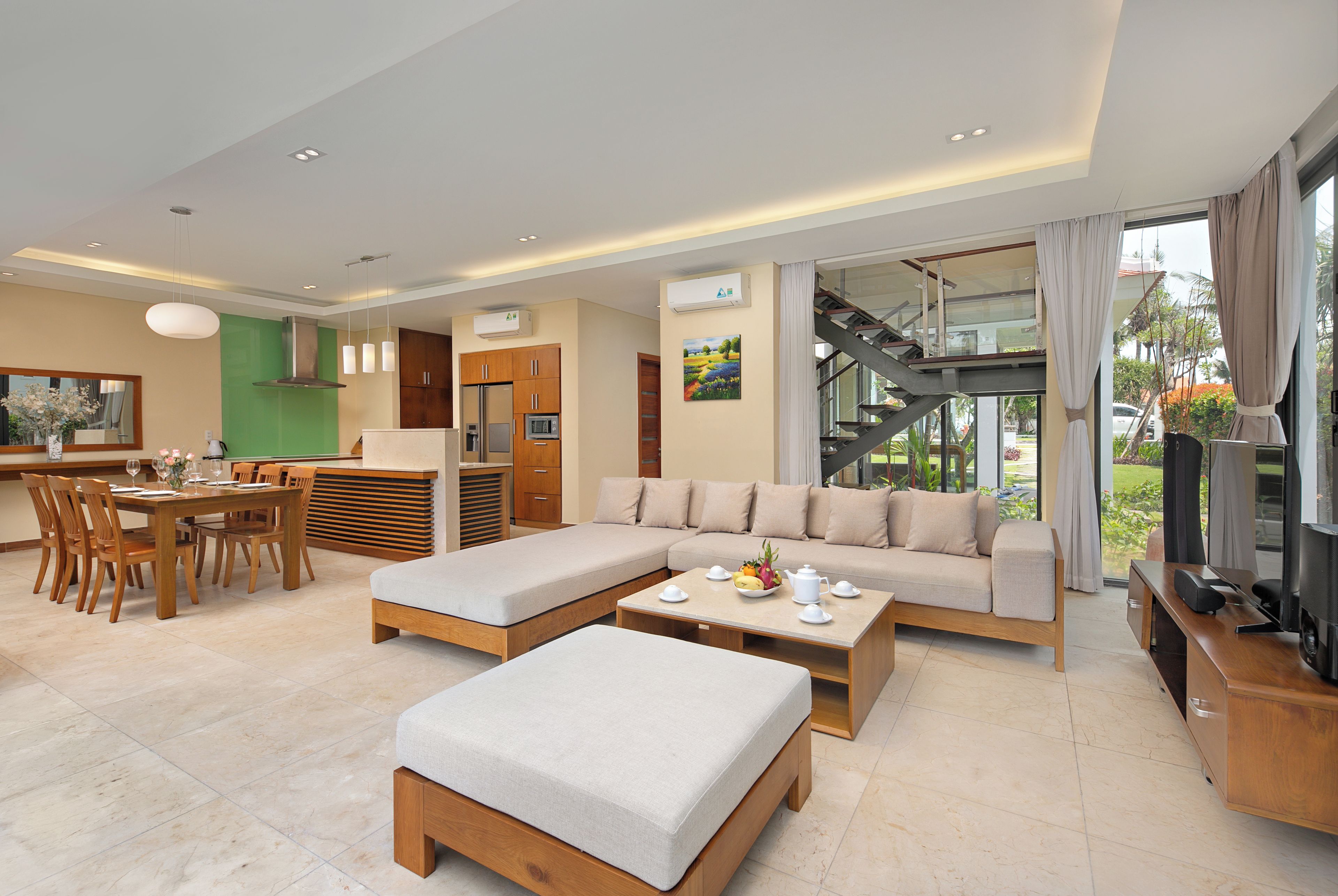 A modern living room with a sofa, ottoman, dining table, and chairs. There is a kitchen area with cabinets and a refrigerator. A staircase leads to an upper level.