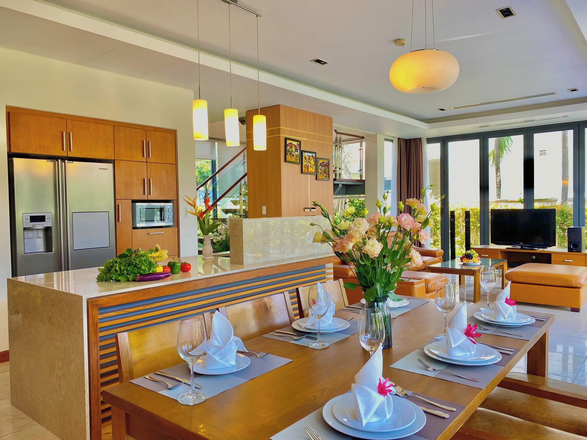 Cocina moderna con electrodomésticos de acero inoxidable, comedor con mesa de madera preparada para cuatro personas y sala de estar con sofá y televisor.