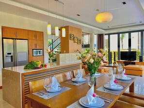 Une cuisine moderne équipée d’appareils électroménagers en acier inoxydable, un coin repas avec une table en bois pour quatre personnes, et un salon avec un canapé et un téléviseur.