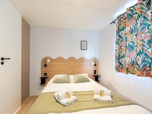 A hotel room with a wooden headboard, two bedside tables with lamps, a bed with green and white bedding, and a colorful curtain. - New Vennepark Caravan 463 (De Haan)