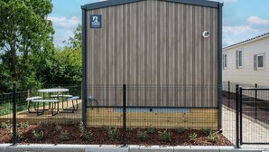 Un edificio de una sola planta con fachada de metal corrugado, un letrero que indica que es un parque para caravanas, y un área exterior cercada con mesas y sillas.