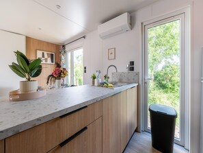 A modern kitchen with a marble countertop, wooden cabinets, a built-in sink, and a large window offering a view of greenery. - New Vennepark Caravan 463 (De Haan)