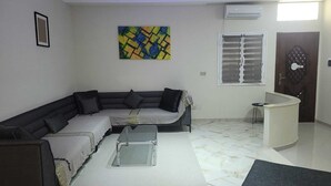Un salon comprenant un canapé d’angle, une table basse, un tapis, un tableau, un climatiseur, une fenêtre avec des stores et une porte décorée d’une guitare.