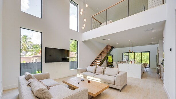 A spacious living area with a large sofa, a coffee table, and a TV mounted on the wall. There is a staircase leading to an upper level with a balcony.