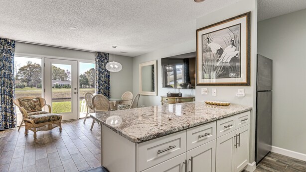 A kitchen with a marble countertop, white cabinets, and a stainless steel refrigerator. There is a dining area with wicker furniture and a view through glass doors. - Water-View Patio! Condo 2 Mi to Surfside Beach (Myrtle Beach)