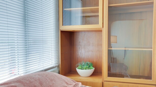 Een kamer met een bed, een raam met zonwering, een houten kastje met een plant en een stoel.
