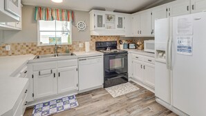 A kitchen with white cabinets, a stove, a refrigerator, and a window with curtains. - Nina's Beach Retreat: ~ 1 Mi to Holden Beach! (Supply)