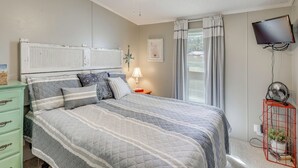 A bedroom with a bed, a ceiling fan, a TV, a nightstand, a dresser, and a window with curtains. - Nina's Beach Retreat: ~ 1 Mi to Holden Beach! (Supply)