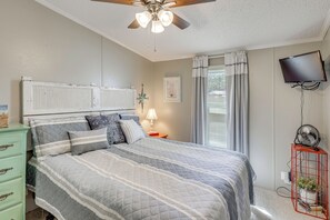 Ein Schlafzimmer mit einem Bett, einem Deckenventilator, einem Fernseher, einem Nachttisch, einer Kommode und einem Fenster mit Vorhängen.