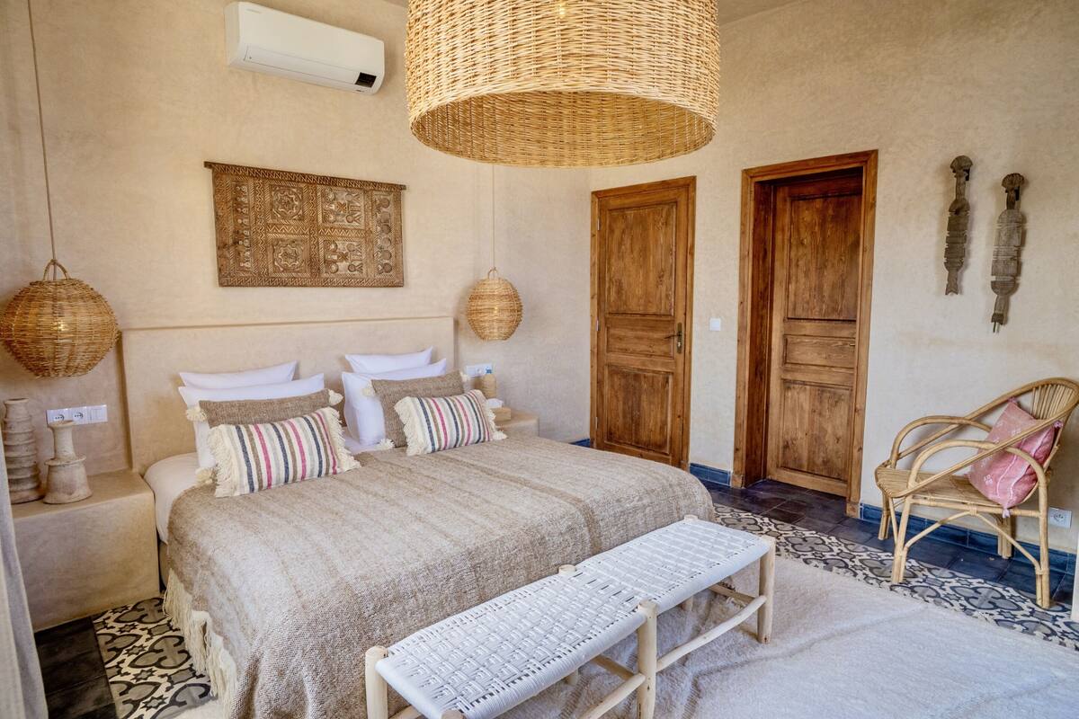 Un dormitorio con una cama grande, dos puertas de madera, lámparas de mimbre, un banco y un tapete estampado.