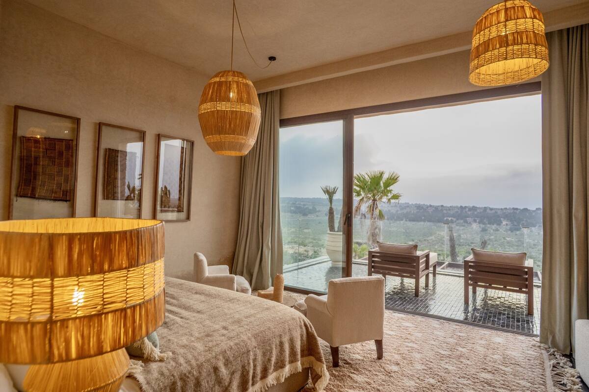 Una habitación de hotel con una cama grande, dos sillas, un balcón con vistas y una lámpara grande.