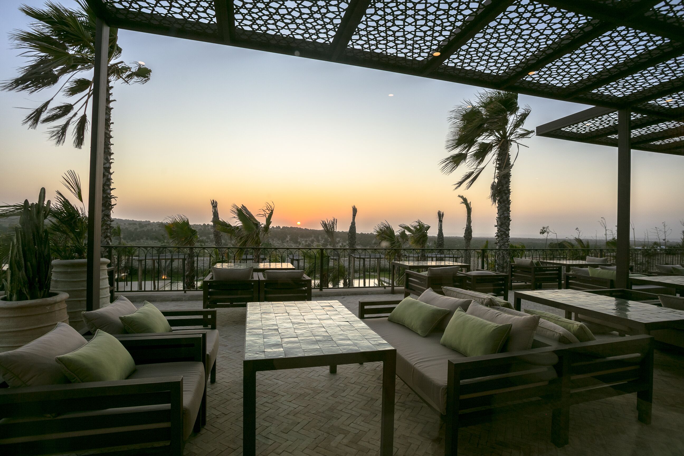 An outdoor seating area with tables and sofas, palm trees, and a view of the sunset.