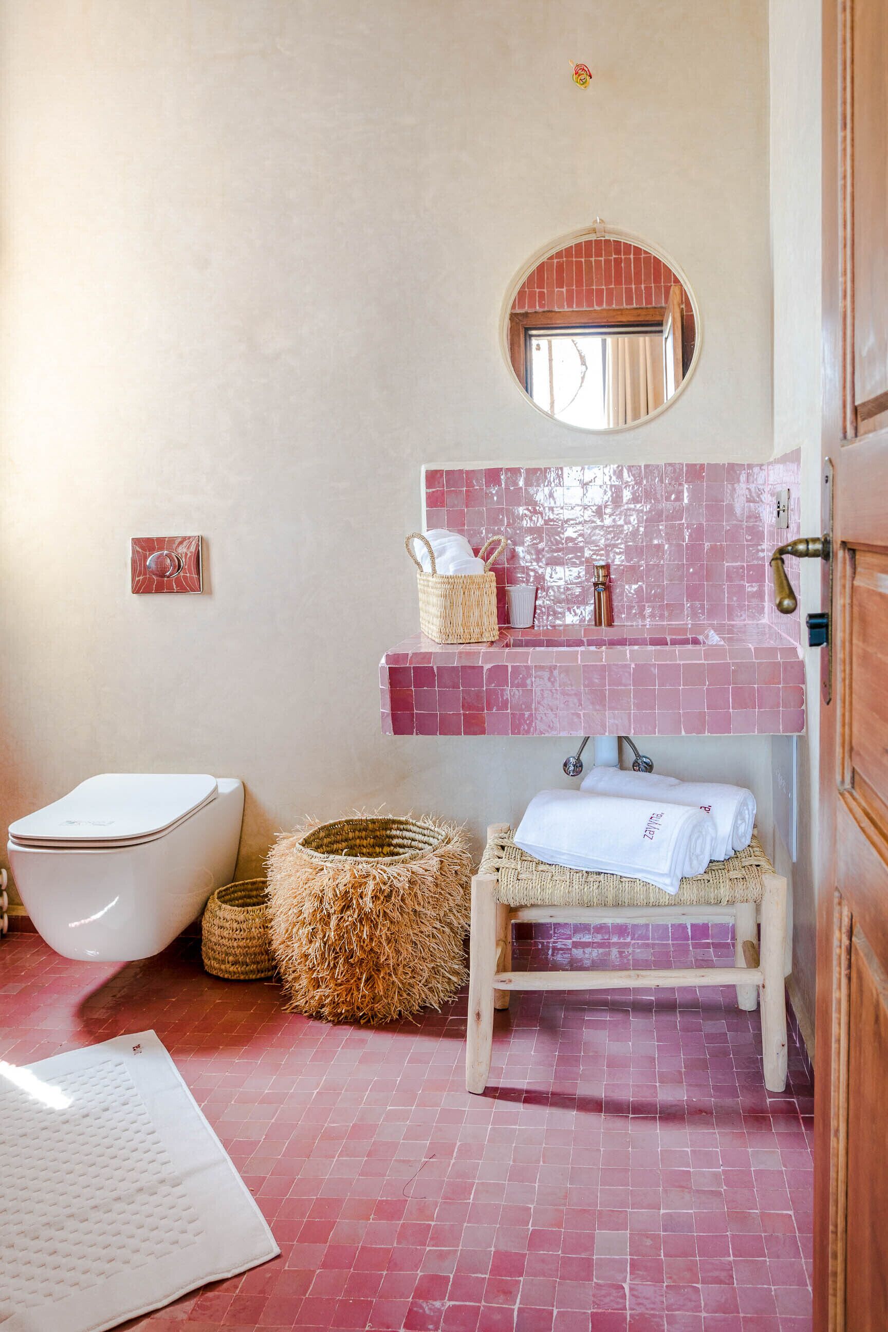 Une salle de bain avec des toilettes blanches, un lavabo avec un pare-éclaboussures carrelé de couleur rose, un miroir et un banc en bois.