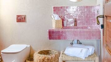 Une salle de bain avec des toilettes blanches, un lavabo avec un pare-éclaboussures carrelé de couleur rose, un miroir et un banc en bois.