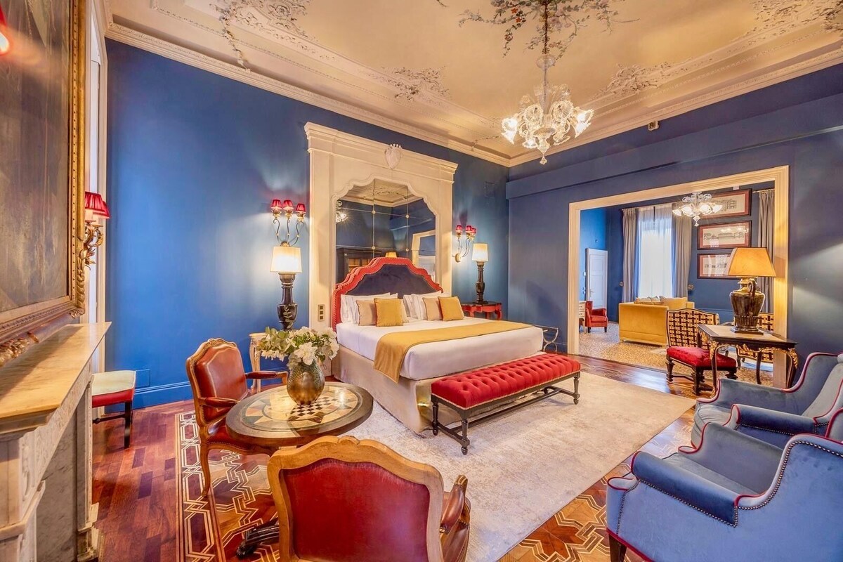 Una lujosa habitación de hotel con una cama grande, un área de estar con sofá y sillones, una mesa de comedor y un candelabro.