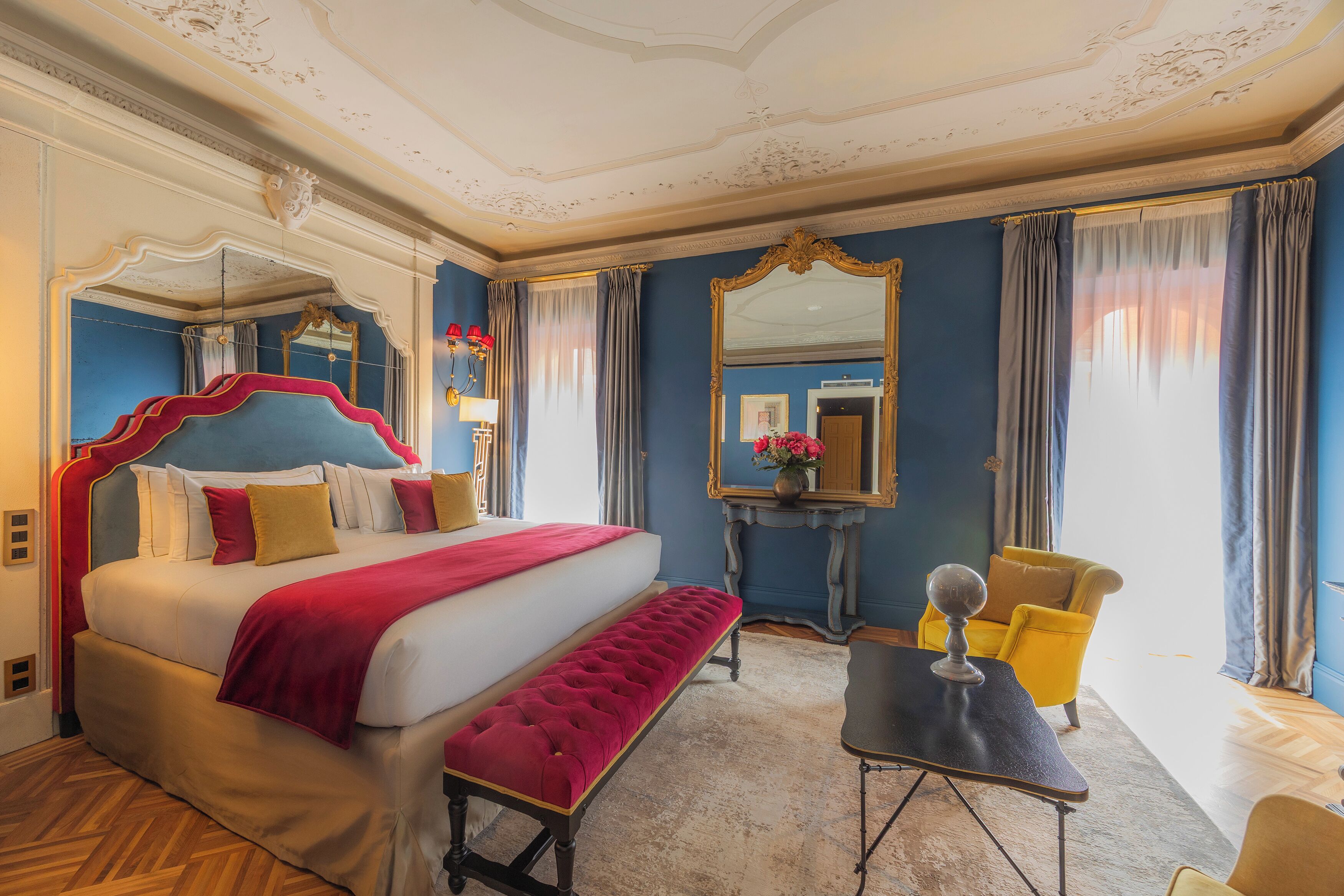 A hotel room with a large bed, a bench, a small table, a chair, and a mirror.