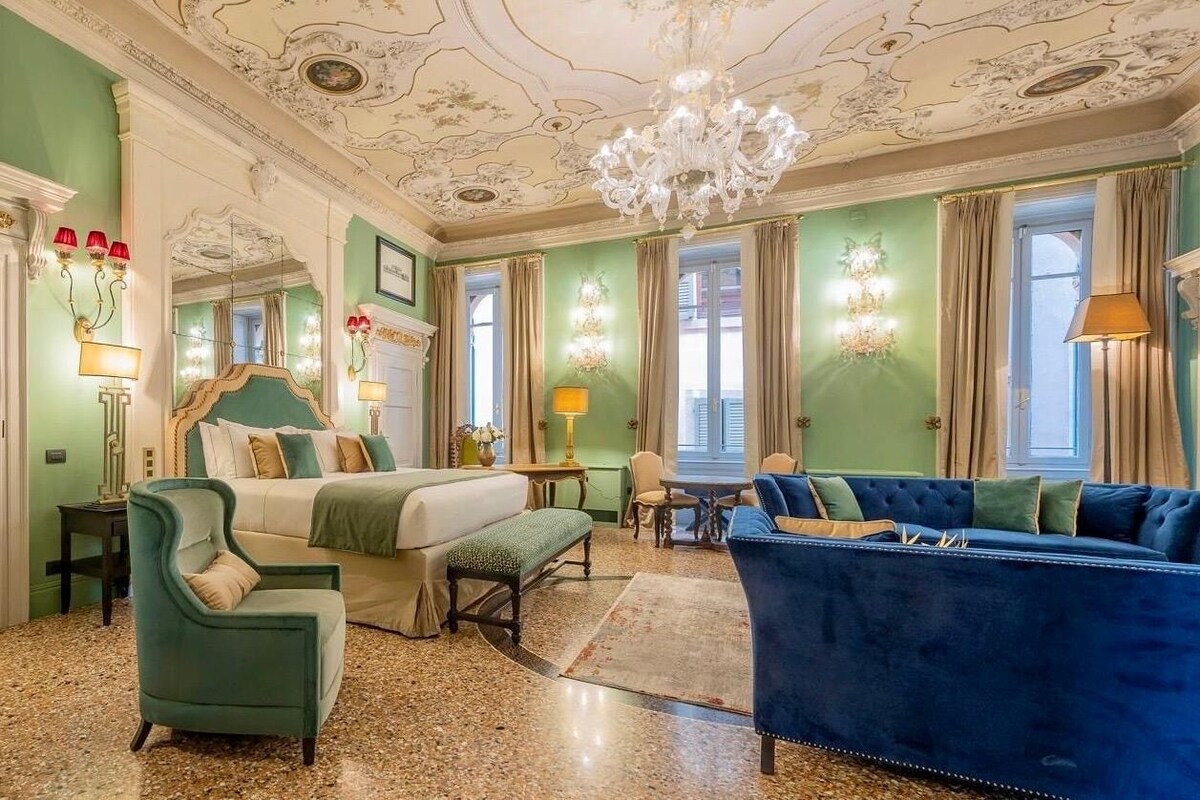 Una habitación con una cama, un sofá azul, un sillón verde, una araña y una mesa de comedor.