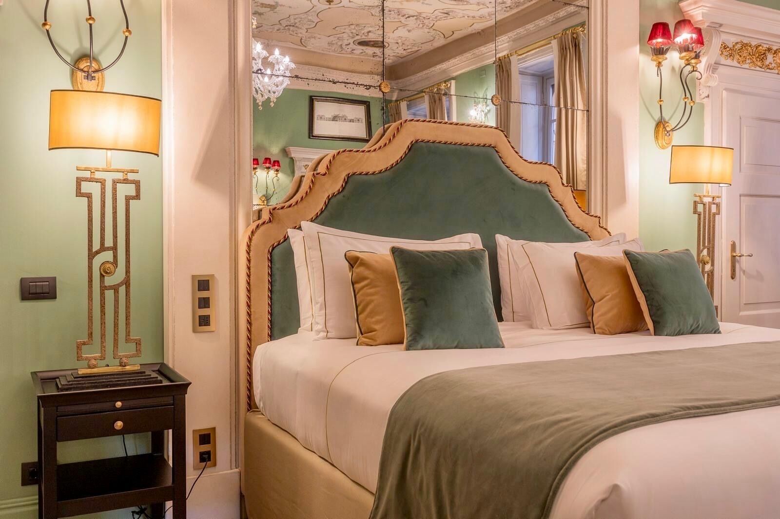 Cama com cabeceira estofada verde, várias almofadas e mesa de cabeceira com abajur.