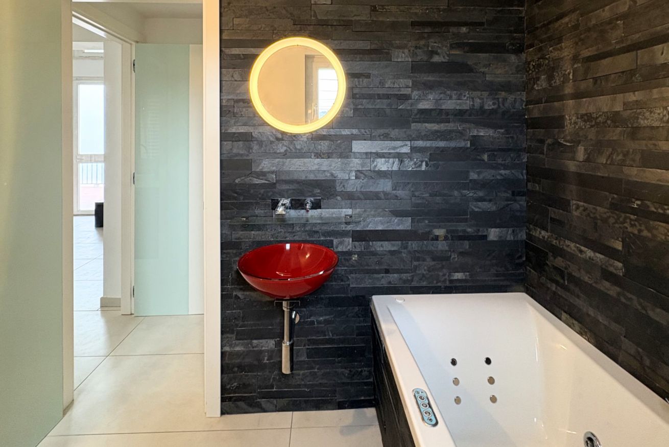 A modern bathroom with a stone wall, a red sink, a white bathtub, and a round mirror.