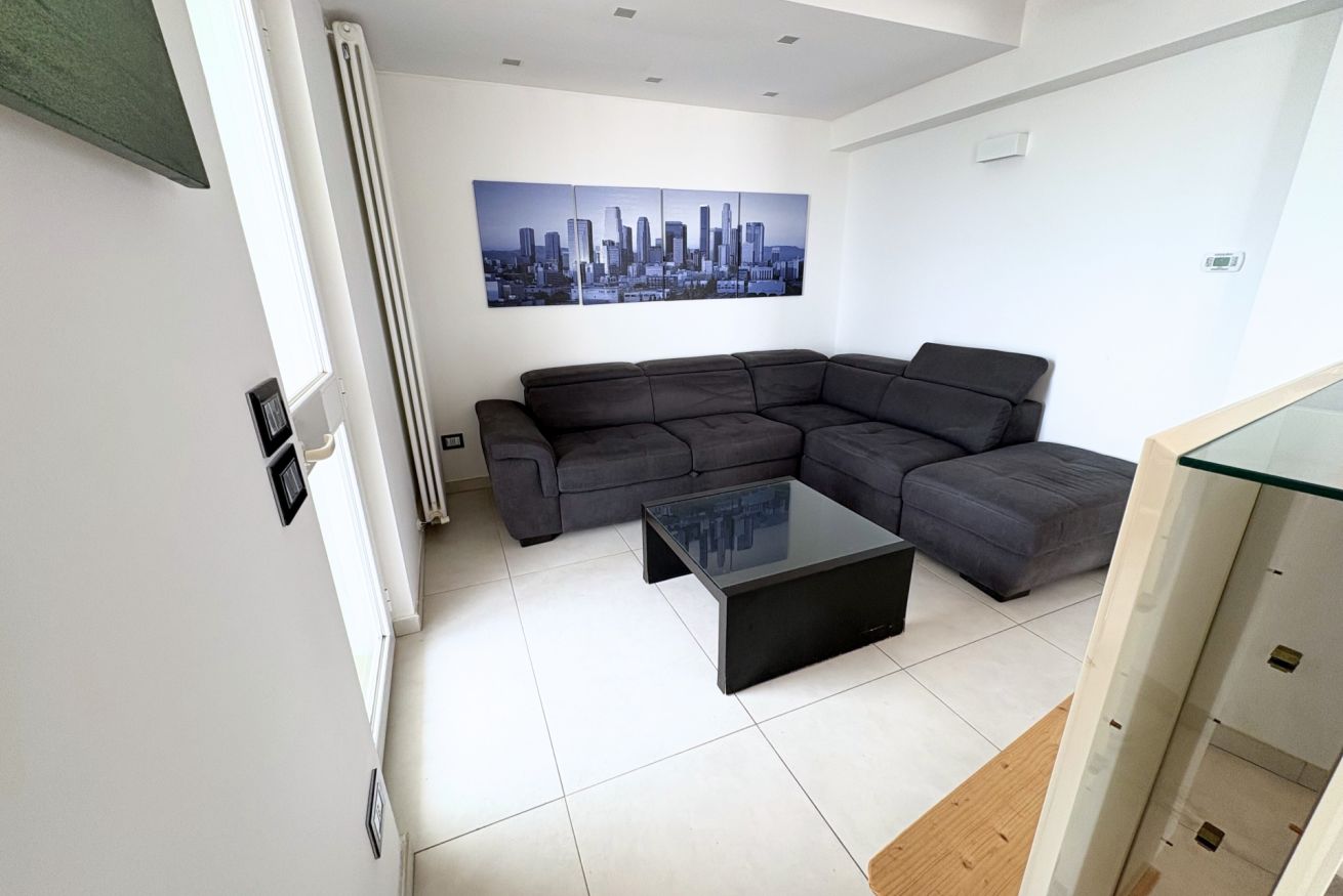 Un salon moderne avec un canapé d’angle, une table basse en verre et un tableau représentant un paysage urbain accroché au mur.