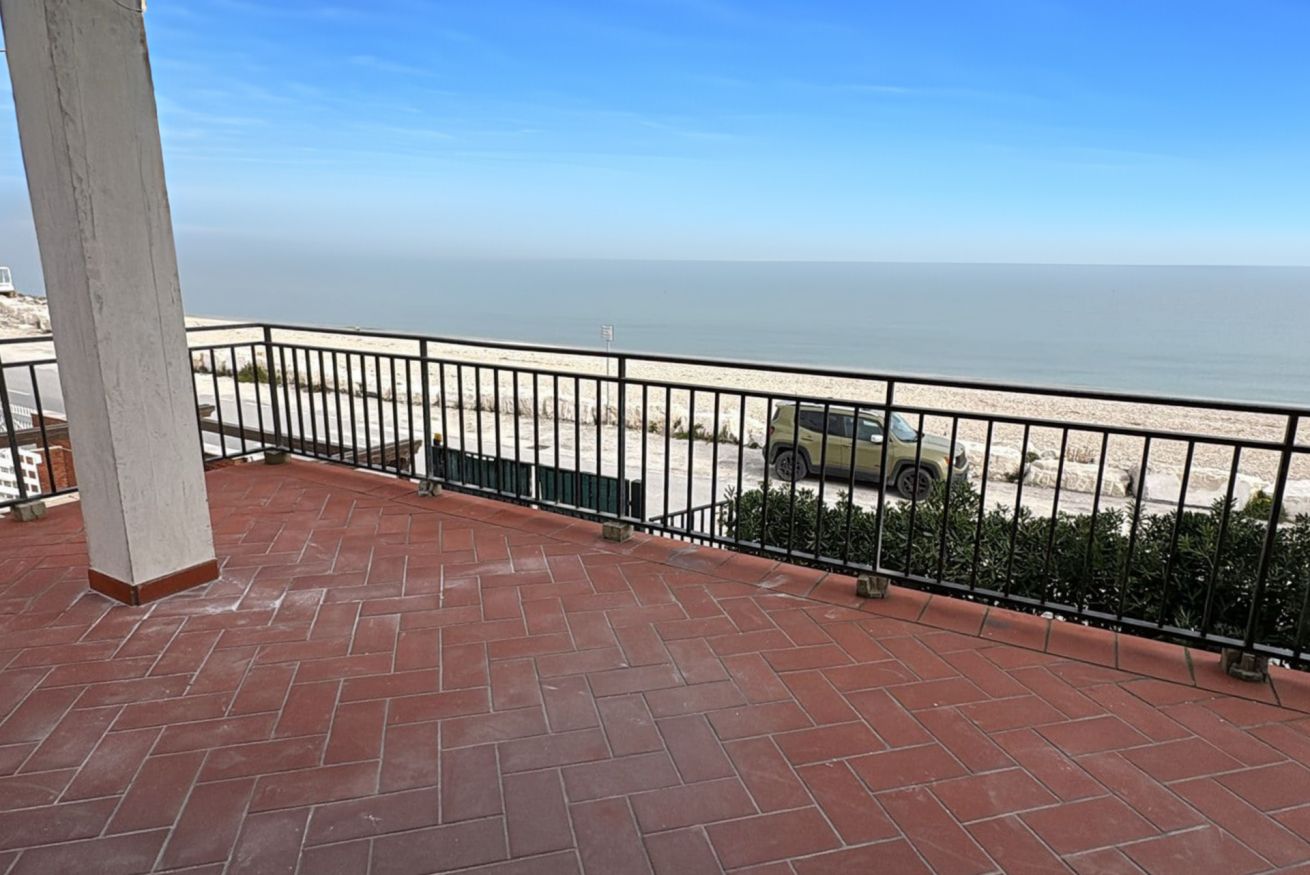 Un balcon avec vue sur la plage et l’océan, un sol pavé et une rambarde en métal noir.