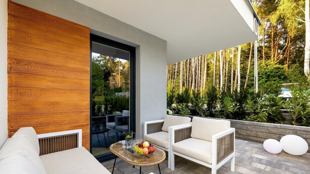 Un espace extérieur moderne, aménagé avec un canapé blanc, une table en osier ornée de fruits et offrant une vue sur une zone boisée.