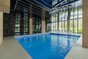 Una piscina interior con paredes de vidrio transparente, suelo de baldosas y vistas a zonas verdes.