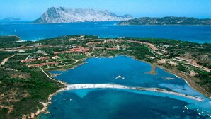 An aerial view of a coastal resort with multiple buildings, a large body of water, and a mountainous backdrop. - Residence Porto Coda Cavallo auf Sardinien (San Teodoro)