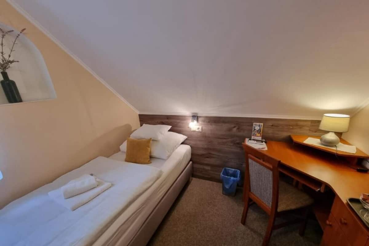 Una habitación pequeña y acogedora con una cama individual, un escritorio con lámpara y un cubo de basura.