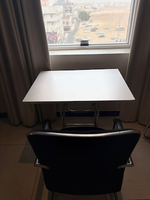 A room with a white table and a black chair, a window with a view of a cityscape, and curtains.