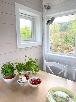 Une pièce avec une table où sont disposés des herbes fraîches, de l’ail et des tomates cerises, une chaise blanche et une fenêtre donnant sur un paysage verdoyant.