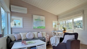 Una sala de estar acogedora con un sofá, un sillón y una mesa de centro con cestas. Hay dos ventanas con cortinas, un cuadro en la pared y un equipo de aire acondicionado.