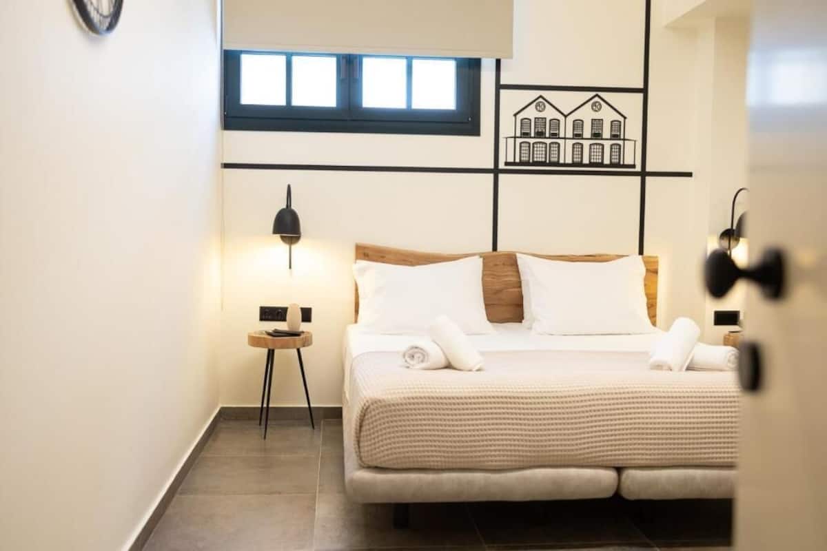 Une chambre à coucher comprenant un lit, des tables de chevet, une lampe, une fenêtre et une œuvre d’art murale.